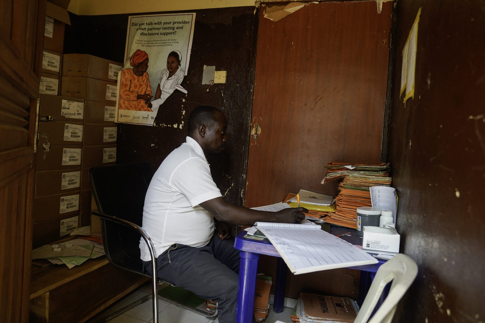 A staff member works at a desk inside the antiretroviral therapy (ART) drug storage room at the Lugbe Primary Health Care Centre in Abuja, Nigeria. Boxes of medical supplies are stacked behind him, and patient records are organized on the desk. July 29, 2025. Photograph: Marvellous Durowaiye /CCIJ